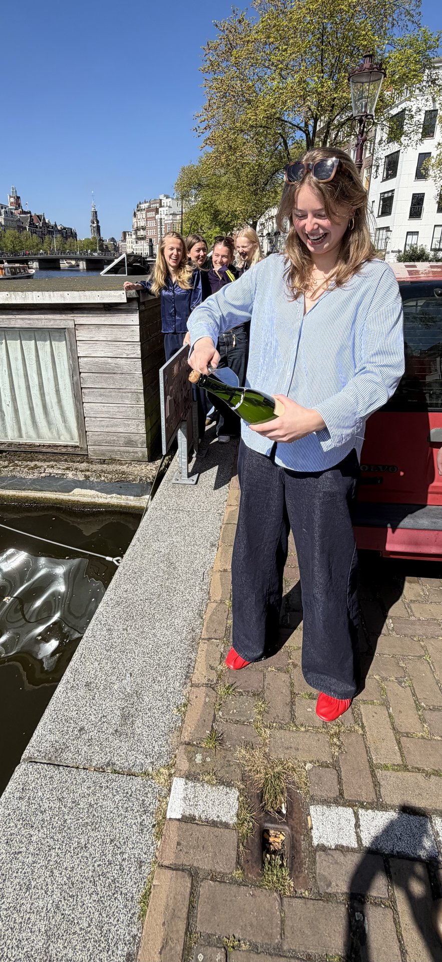 A group of friends enjoys a sunny day at Staalkade, Centrum, Amsterdam, as one person joyfully opens a bottle of sparkling wine by the canal